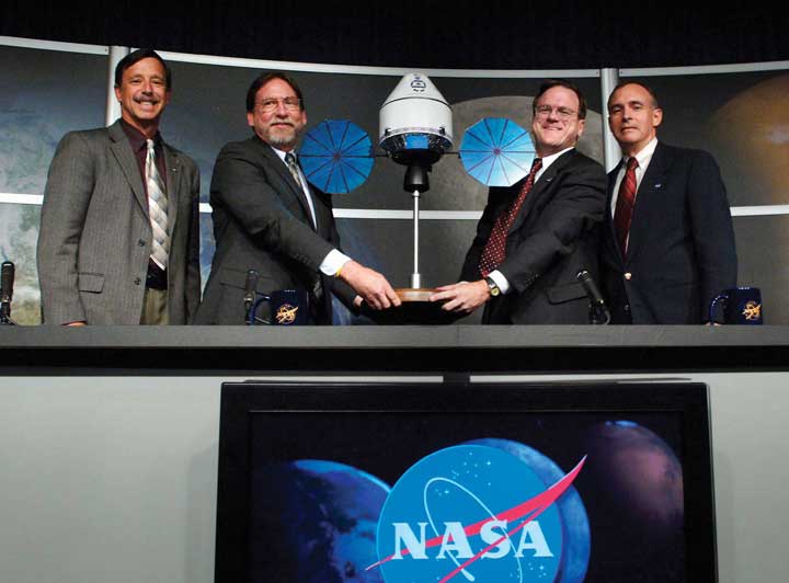 Four men at a table showing off a model of the NASA Orion module