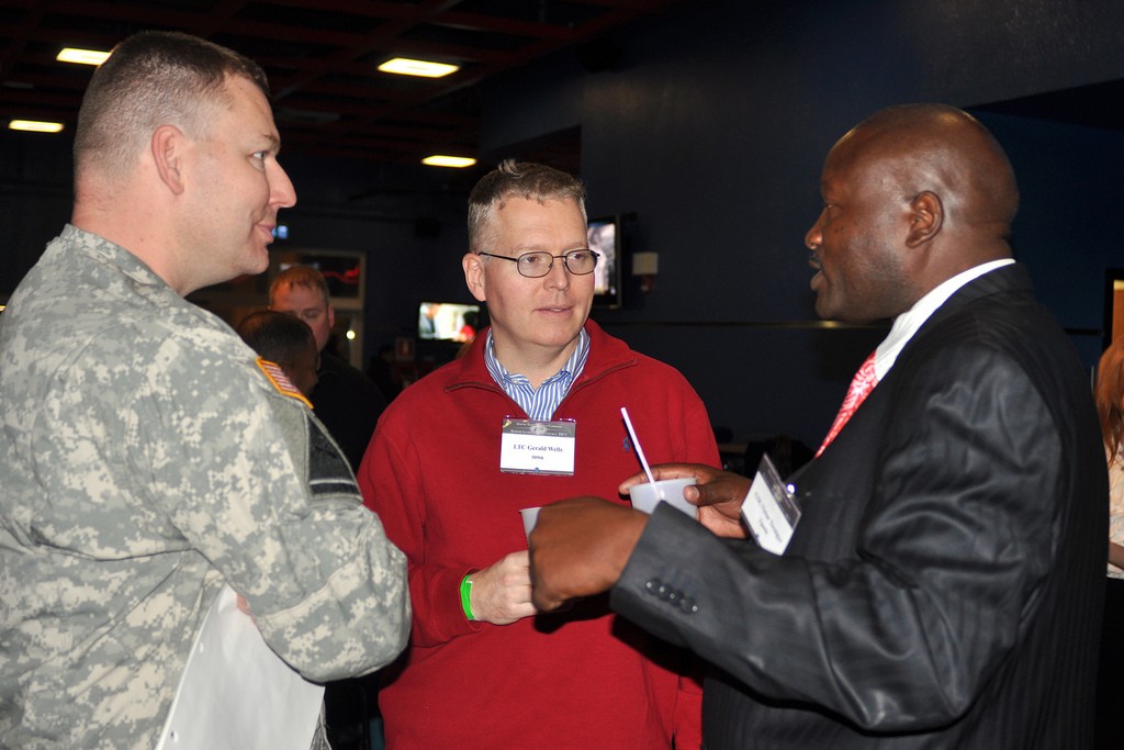 Three men talking, one in fatigues, one in informal sweater, one in business dress