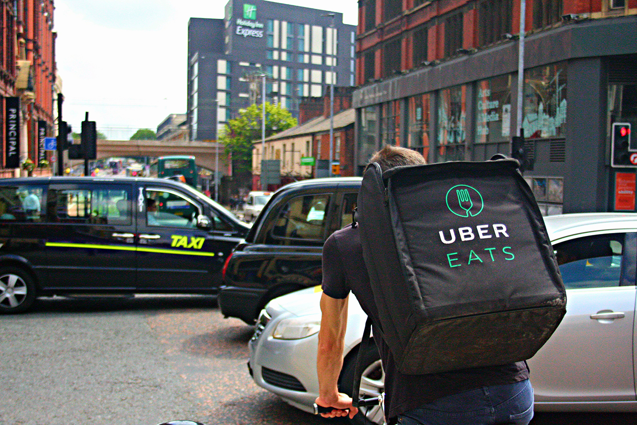 صورة تُظهر المنظر الخلفي لسائق توصيل الطعام في أوبر إيتس مع حقيبة ظهر، وهو يقود دراجة على طول طريق أكسفورد المزدحم في مانشستر، إنجلترا.