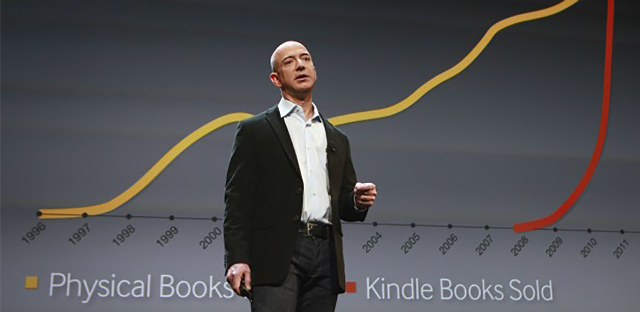 Uma foto mostra Jeff Bezos exibindo o slide mostrando o crescimento fenomenal das vendas de e-books Kindle da Amazon em comparação com as vendas de livros físicos durante a apresentação dos novos Kindles.