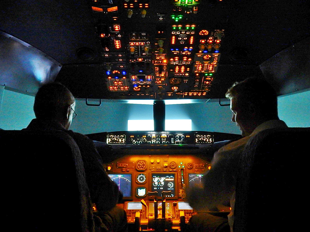 Uma foto mostra a visão traseira de um instrutor de voo descrevendo vários mostradores e displays para um piloto estagiário na cabine de um simulador de Boeing 7 3 7.