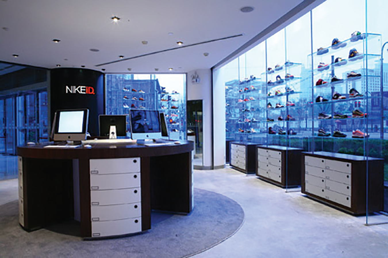 Une photo de l'intérieur d'un magasin NikeID montrant des chaussures sur des étagères près de fenêtres vitrées avec vue sur de nombreux bâtiments de grande hauteur.