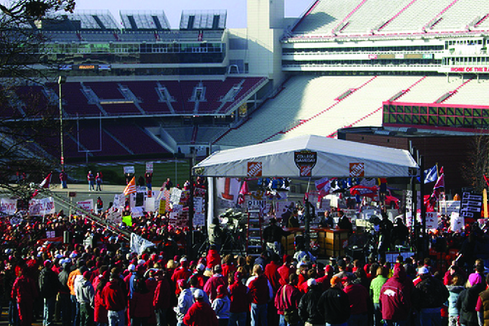 A photograph shows a crowd of people standing near a stadium at a tent.