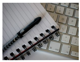 Imagen de un bolígrafo negro encima de un cuaderno de espiral abierto encima de un teclado blanco.