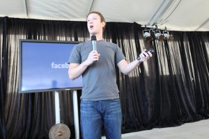A photograph of Mark Zuckerberg, dressed in jeans and t-shirt, holding a microphone and giving a talk in front of a small screen with a blue background that reads “Facebook.”