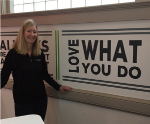 A photograph of Amy Ankrum, standing to the left of a wall that reads “Love What You Do” Where love is written sideways.