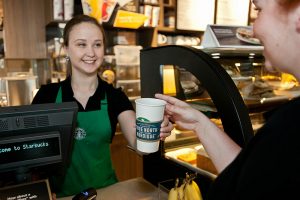 A photograph of a Starbucks barista behind a counter in Starbucks, serving a coffee to a customer while smiling.