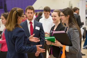 A photograph of a recruiter talking to three students at a college campus job fair. They are all dressed in business professional clothing.