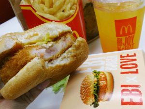 A photograph of a McDonald’s meal from Singapore. An order of fries and an orange drink are in the background. A prawn burger is in the foreground with a bite out of it, along with the box it came in to the right.