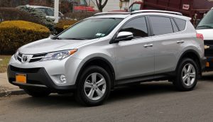 A photograph of a silver RAV-4, shown from the side. It is parallel parked on a street, with another car behind it and bushes in the background.