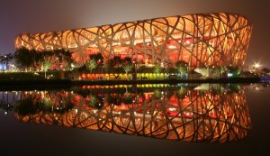 A photograph of the Beijing National Stadium at night, taken from across water. The National Stadium is lit up by yellow lights from within. A reflection of the stadium appears on the water.