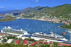 A photograph of two cruise ships docked in a bay, Charlotte Amalie, in the Virgin Islands. On land beside the cruise ships are eight buildings with red roofs. Smaller boats are in the bay away from the cruise ships. A mountain range lies in the background.