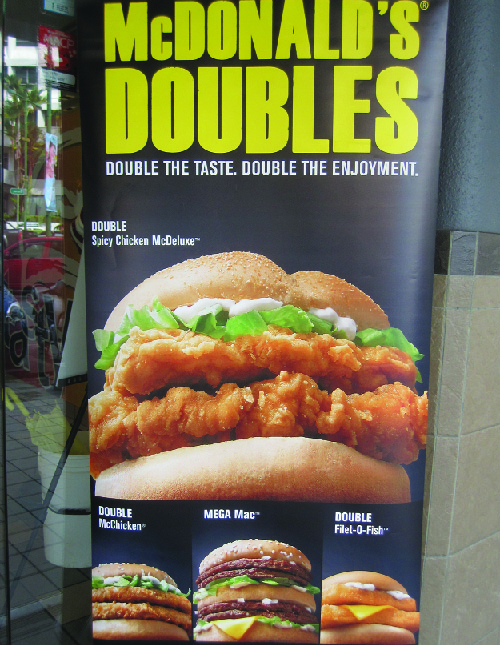 这张照片显示了麦当劳双打的广告。 上面写着 “味道加倍。 享受双倍。” 它显示了四个三明治：Double Spicy Chicken mcDeluxe、Double McChicken、Mega Mac 和 Double Filet-O-Fis