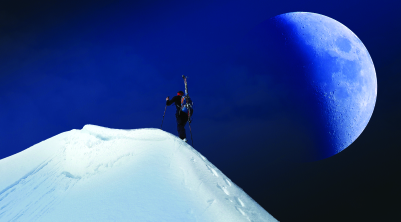 这张照片显示了一个人在积雪覆盖的山上徒步旅行。 月亮已经半满了，在山和徒步旅行者的右边。 它在天空中看起来很大，离徒步旅行者很近。