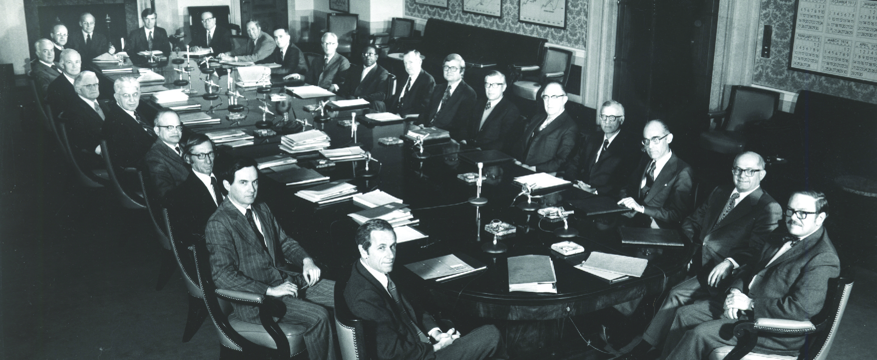 Esta imagem mostra vinte e três homens brancos e um homem negro em ternos sentados ao redor de uma grande mesa em estilo de sala de reuniões.