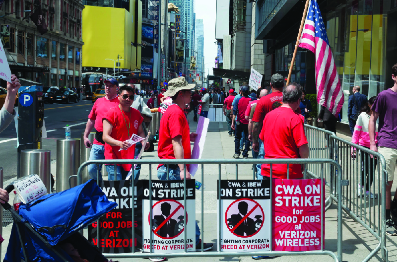 Esta imagem mostra um grupo de pessoas vestidas com camisas vermelhas combinando em uma área cercada por placas que dizem “CWA em greve por bons empregos na Verizon Wireless” e “CWA e IBEW em greve. Combatendo a ganância corporativa na Verizon.”