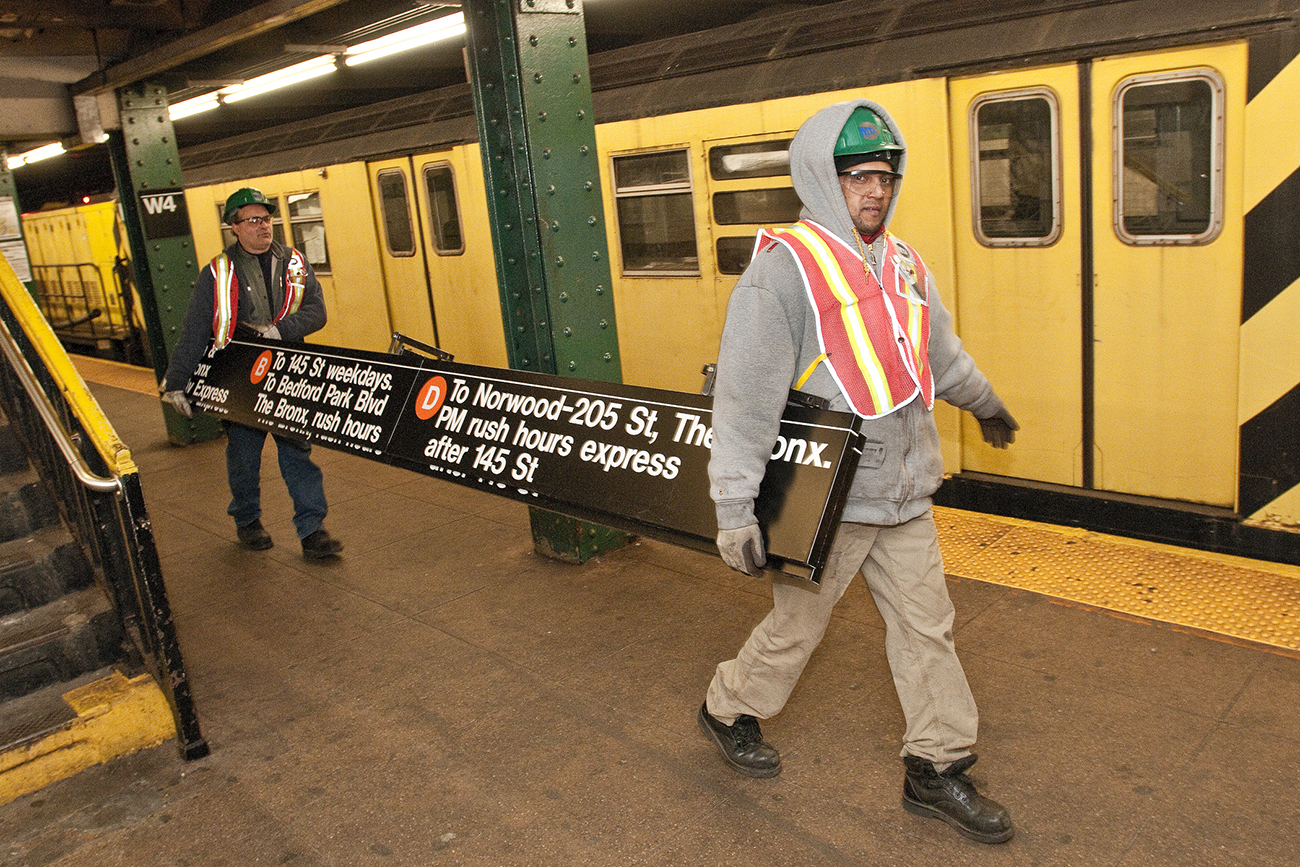 Une photo montre deux employés du métro au travail. Ils portent un panneau de destination à l'intérieur d'une station de métro.