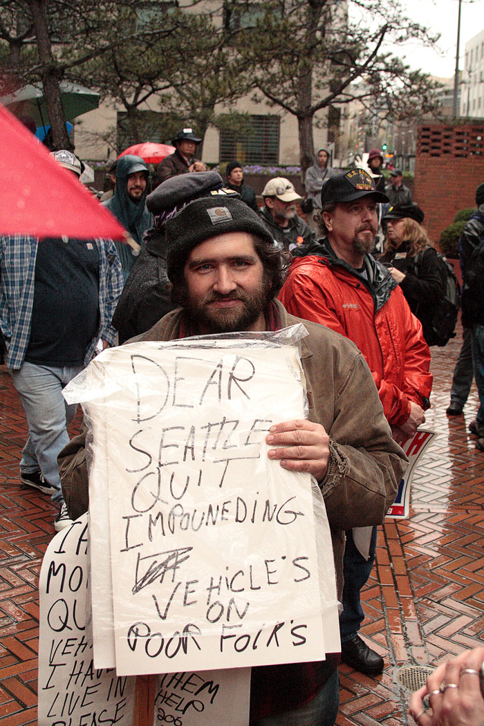 Une photo montre un manifestant lors d'un rassemblement de protestation contre la guerre, tenant une pancarte sur laquelle on peut lire : « Chère Seattle, arrête de saisir des véhicules sur les pauvres ».