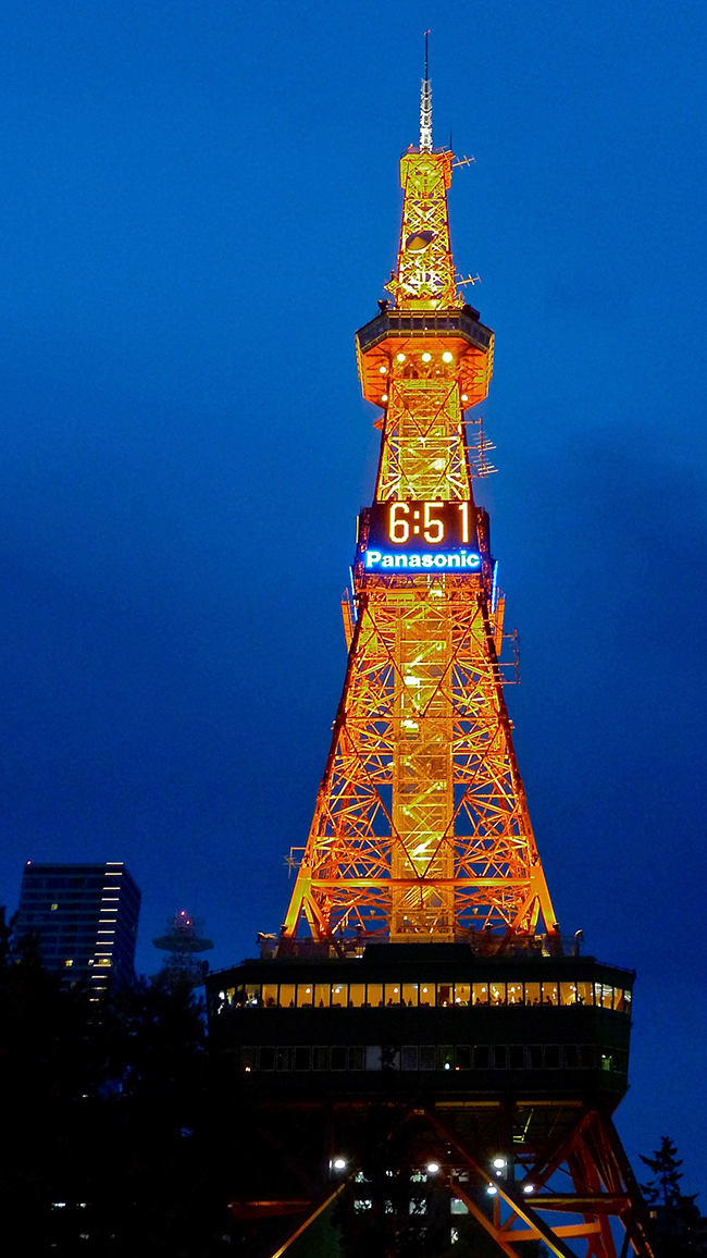 A photo of the Sapporo TV tower with digital clocks installed on it at four sides. The digital clocks read “6:51” with the advertisement of Panasonic right below the clocks.