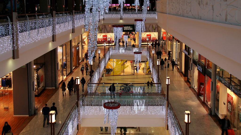 Photograph of a shopping mall taken from the third floor of the mall, and the second and first floor can be seen. The center of the mall is open and bridged with walk ways. The mall is decorated for winter, and lights hang from the balconies of the third and second floors.