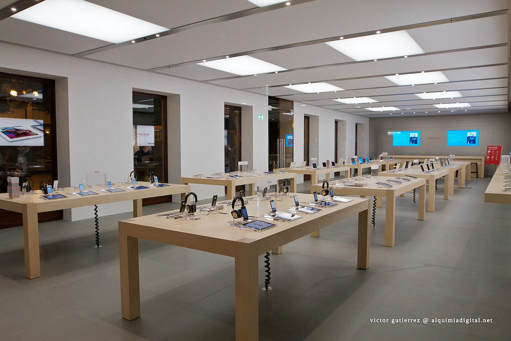 An apple store layout. There are two rows of three tables with a few items displayed on each surface.