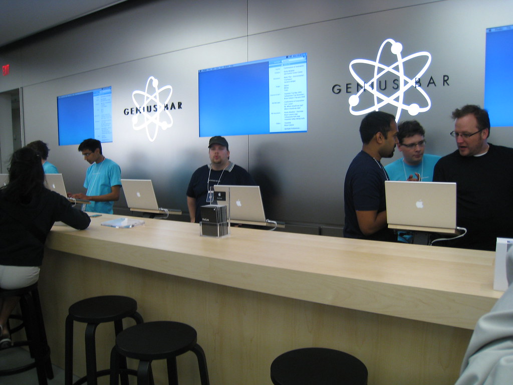 The genius bar at an apple store. Several employees sit behind a counter at Apple computers.
