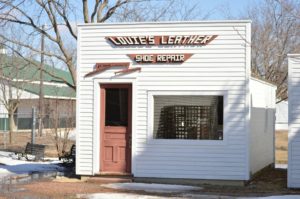 A storefront called "Louie's Leather Shoe Repair". The building is white with a brown door. There is another building in the background and some trees. The trees are bare and there is snow on the ground.