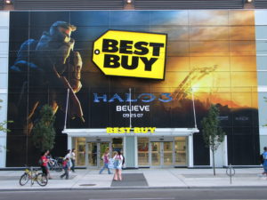 Image of a Best Buy storefront with people walking along the sidewalk that runs in front of the entrance doors.