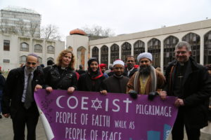 Photo of six people of different religious holding a purple poster with white text. The sign says "Coexist pilgrimage people of faith walk as people of peace".