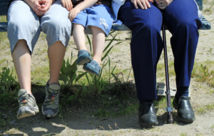 Three people of different ages sitting on a bench. The person on the left is in the 30-40 year-old range, the person in the middle is in the 5-10 year old range, and the person on the left has a cane and is in the 60-75 year old range.