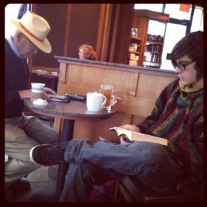 Two men of different ages sitting at a table at a coffee shop. On the left, an older man is reading from a Kindle, while on the right, a young man is reading a book.