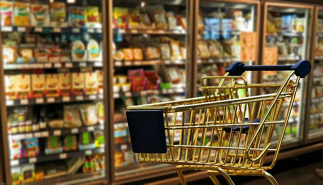 A golden shopping cart is stationary in front of the frozen foods aisle at a grocery store. The shopping cart is empty.