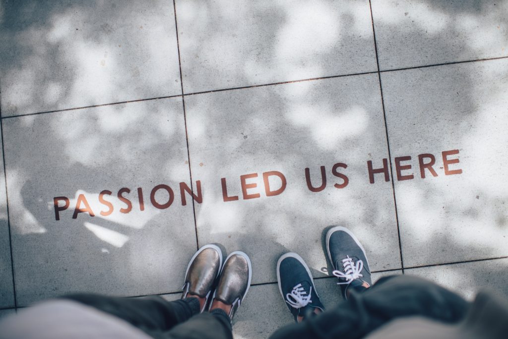 Aerial photograph of two people standing on the sidewalk. They are only shown from the hips down. On the sidewalk are the words "Passion led us here."
