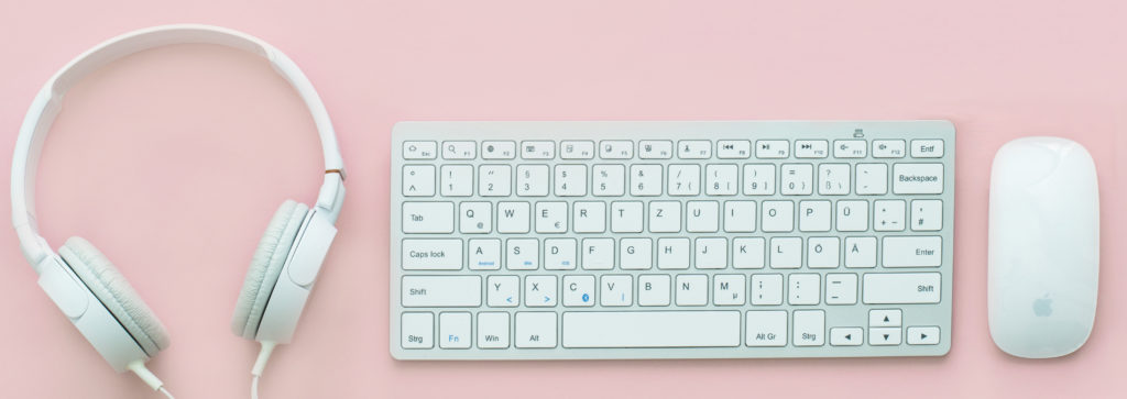 Photograph of a set of headphones, a keyboard, and a mouse on a pink background.