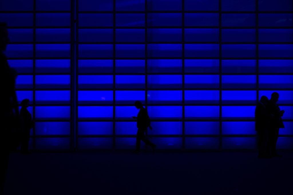 Photograph of a dark room with a blue lit wall. There are shadows of people walking in front of it.