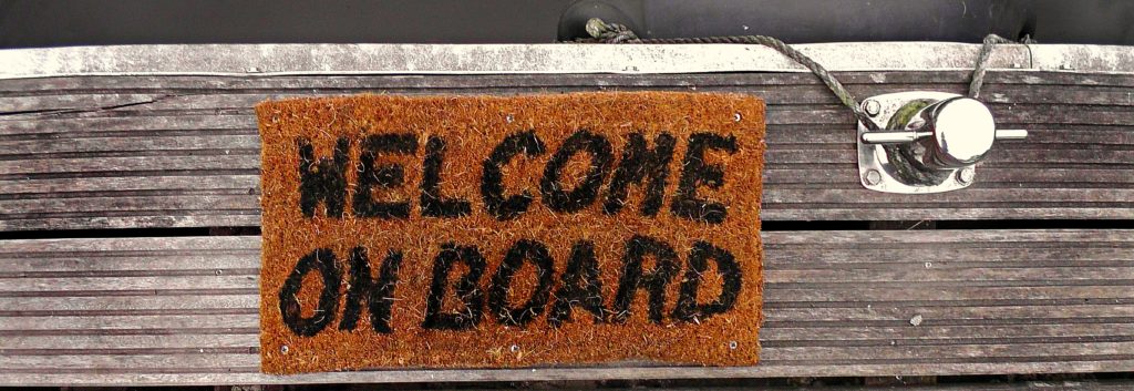Photograph of a welcome mat on a ship dock. The mat reads "Welcome on board."