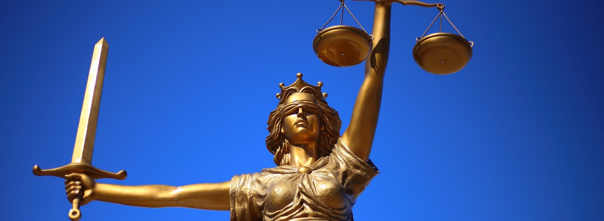 Cropped image a of a statue of justice personified as a woman. In one hand she is holding a sword, in the other she is holding scales. She is also blindfolded.