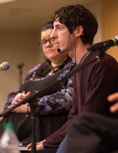 A side view of Jame Damore sitting on a stage with a microphone. Helen Pluckrose is sitting in the background watching Damore speak.