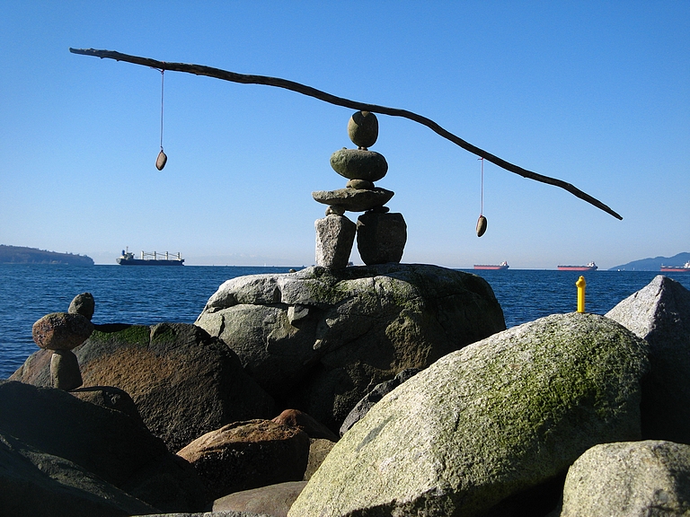 A scenic photograph of a rocky, cliffy area looking out into the water. On the cliff, some rocks are stacked to make a cairn, and on top of the cairn is a stick with two small rocks on either side. The stick is balanced carefully like a teeter-totter on the rocks.