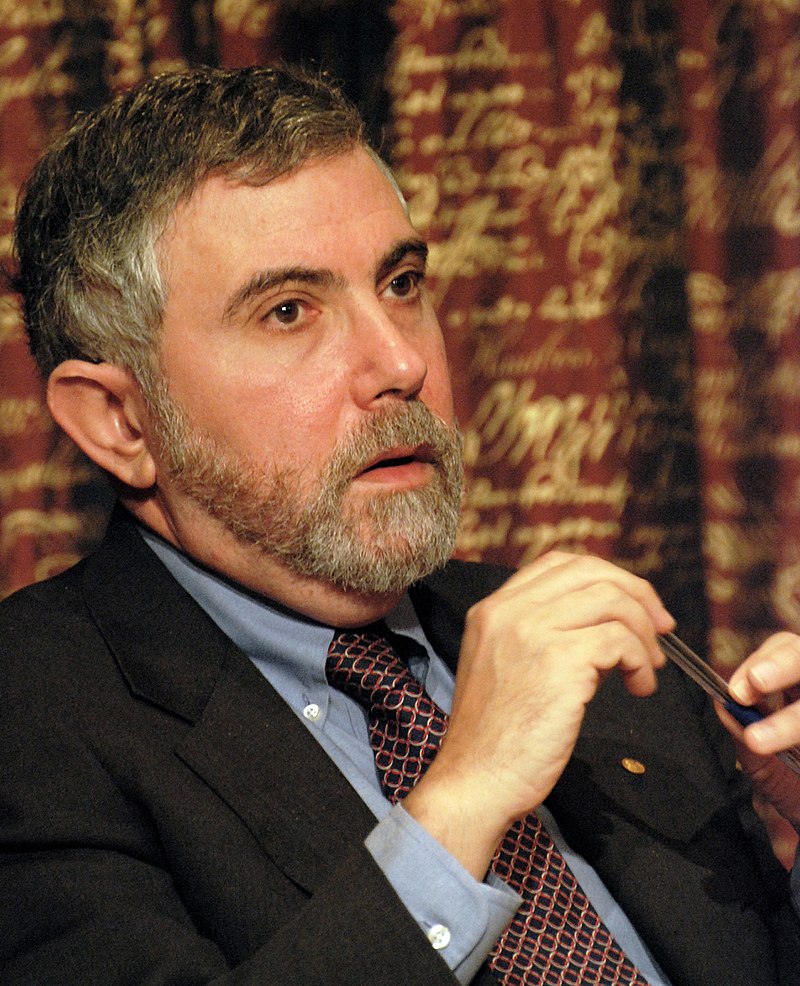 Image of Paul Krugman, sitting, wearing a suit. He's white, with a peppered gray hair and a beard.