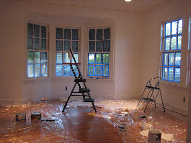 A white-walled room with two step stools, clear plastic, and several cans of paint on the floor