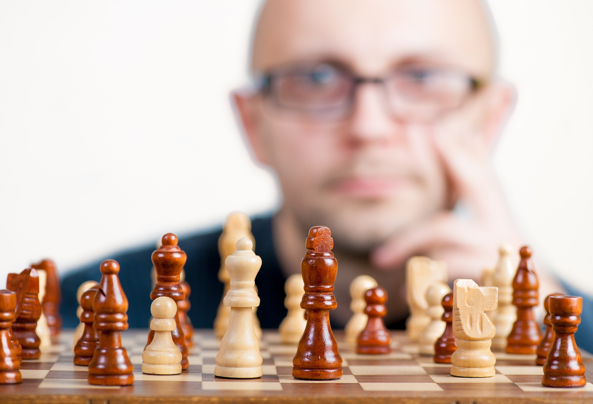 Pieces set up on a chess board, with a man appearing out of focus in the background with a thoughtful expression on his face