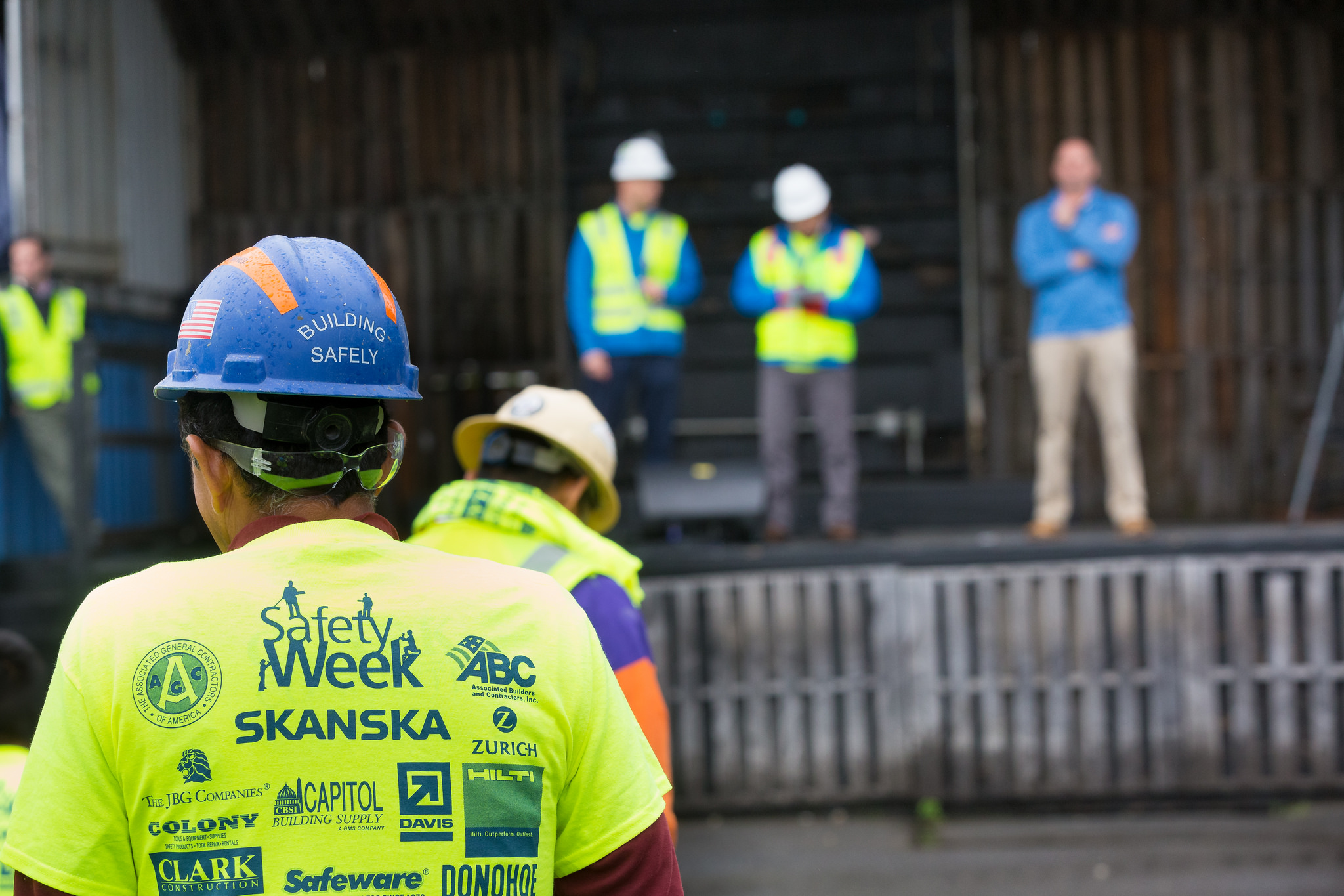 In the foreground, there's a worker wearing a hard hat and a yellow shirt that reads Safety Week. In the background, there are workers wearing hard hats and safety vests standing in an unfinished building.