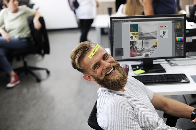 A male employee sitting in front of a computer, dressed casually and smiling at the camera. He has a sticker on his forehead that reads “Be Happy.”