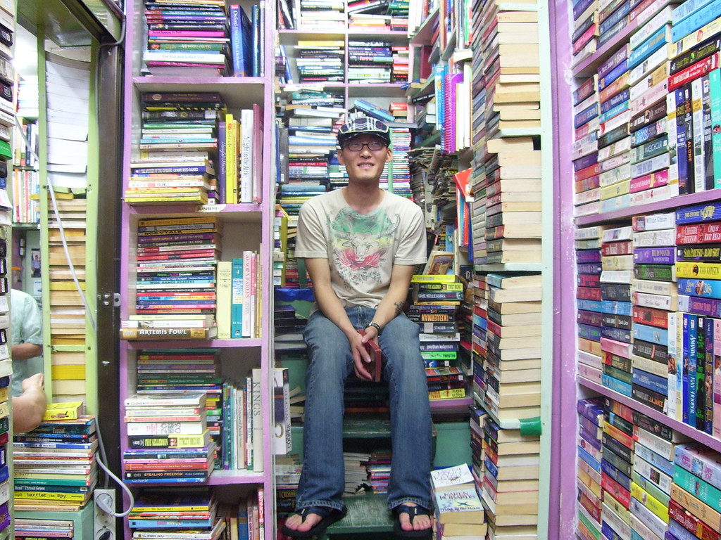A man sits in the middle of a book store with books stacked from floor to ceiling on shelves. The man is wearing a hat and flip-flops and looking around at the books.