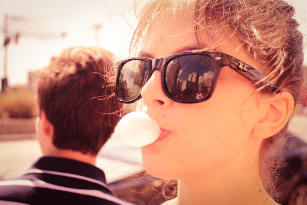 Teenage girl in foreground wearing sunglasses, blowing a gum bubble. Boy in background.