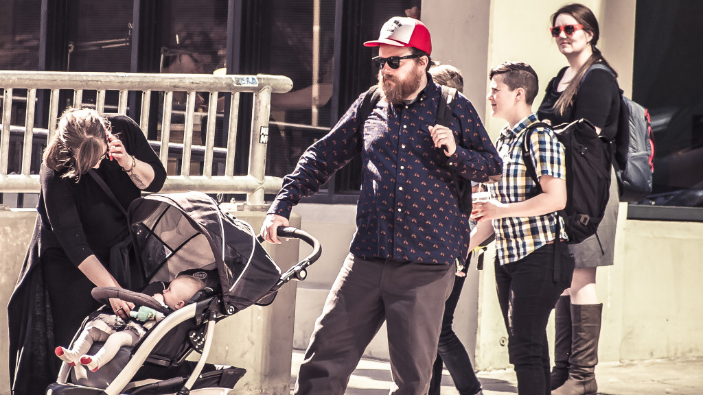 Guy with a beard wearing a red hat pushes a stroller while a woman checks the child and talks on her cell phone. Two young people in the background. Seattle hipsters.