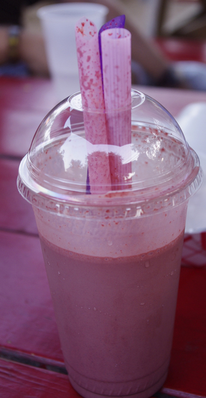 Photo of a pink-colored milkshake in a plastic cup with a plastic lid and three large straws.