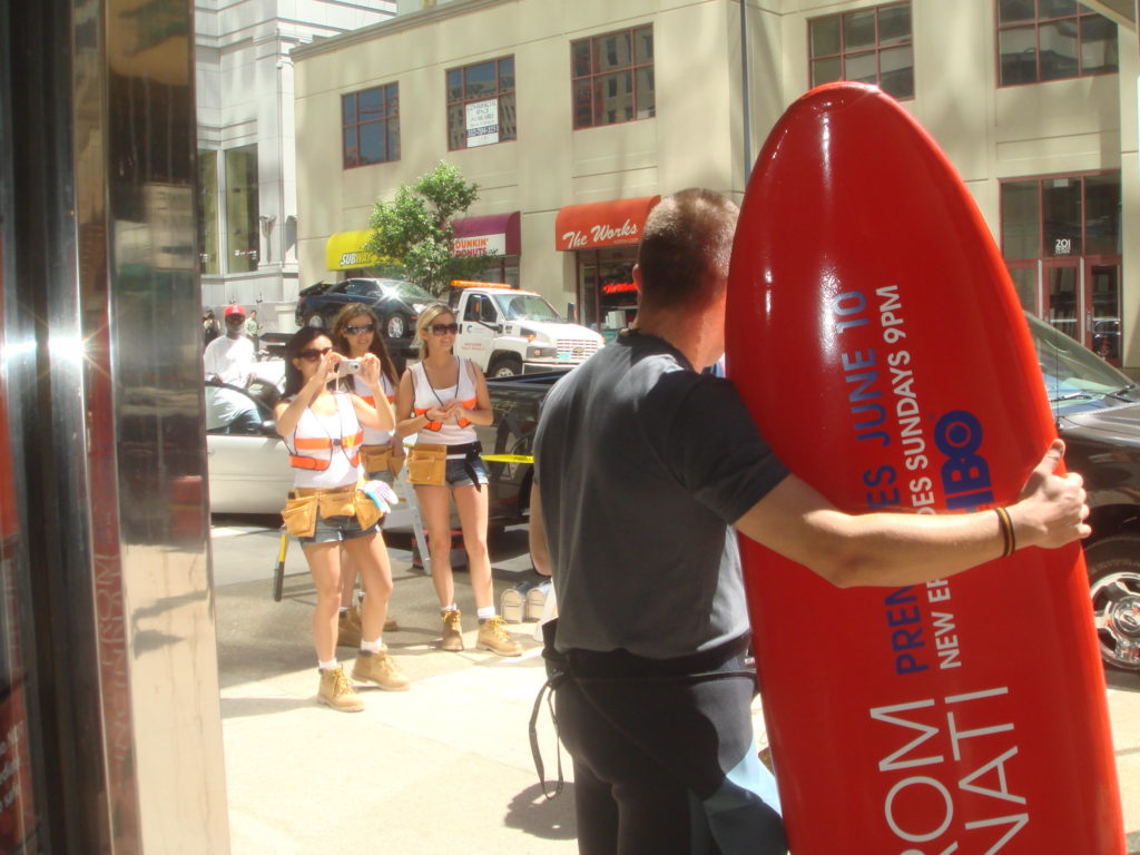 Photograph of a man with a bright red surfboard with an ad for "John from Cincinnati," an HBO show. There are Women wearing safety vests in the background of the photo.