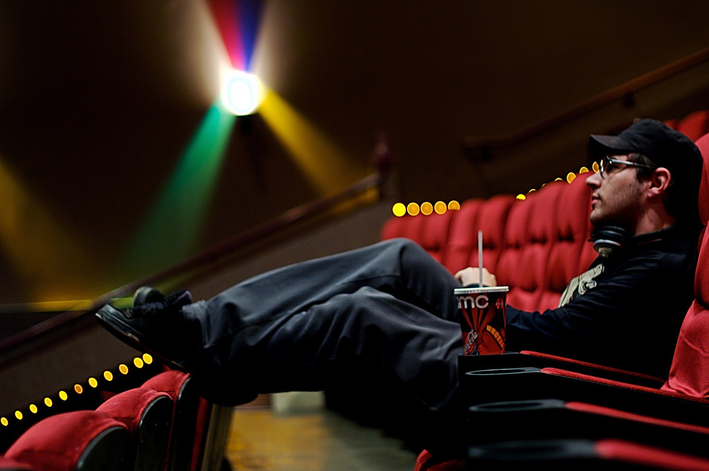 Photo of a man wearing sunglasses, sitting in an empty movie theater holding a soda.